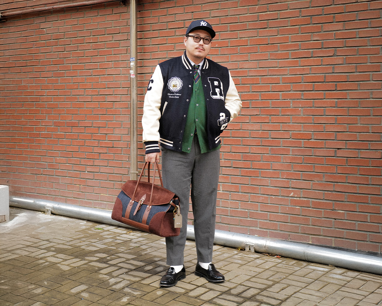 a man wears varsity jacket and new york yankees cap and grey trousers