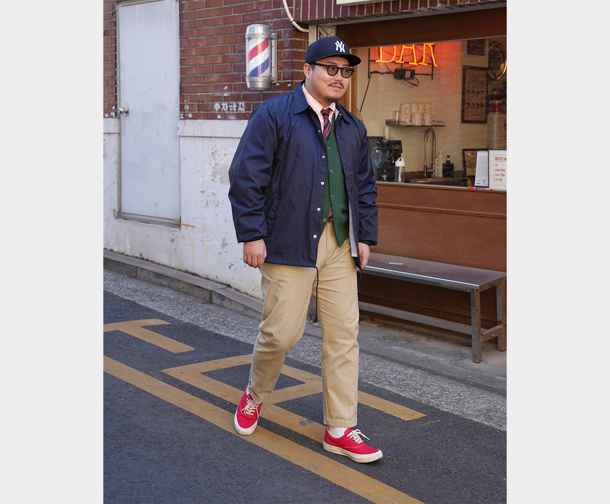 A street style photo of a man wearing a navy coach jacket, green cardigan, and beige chinos.