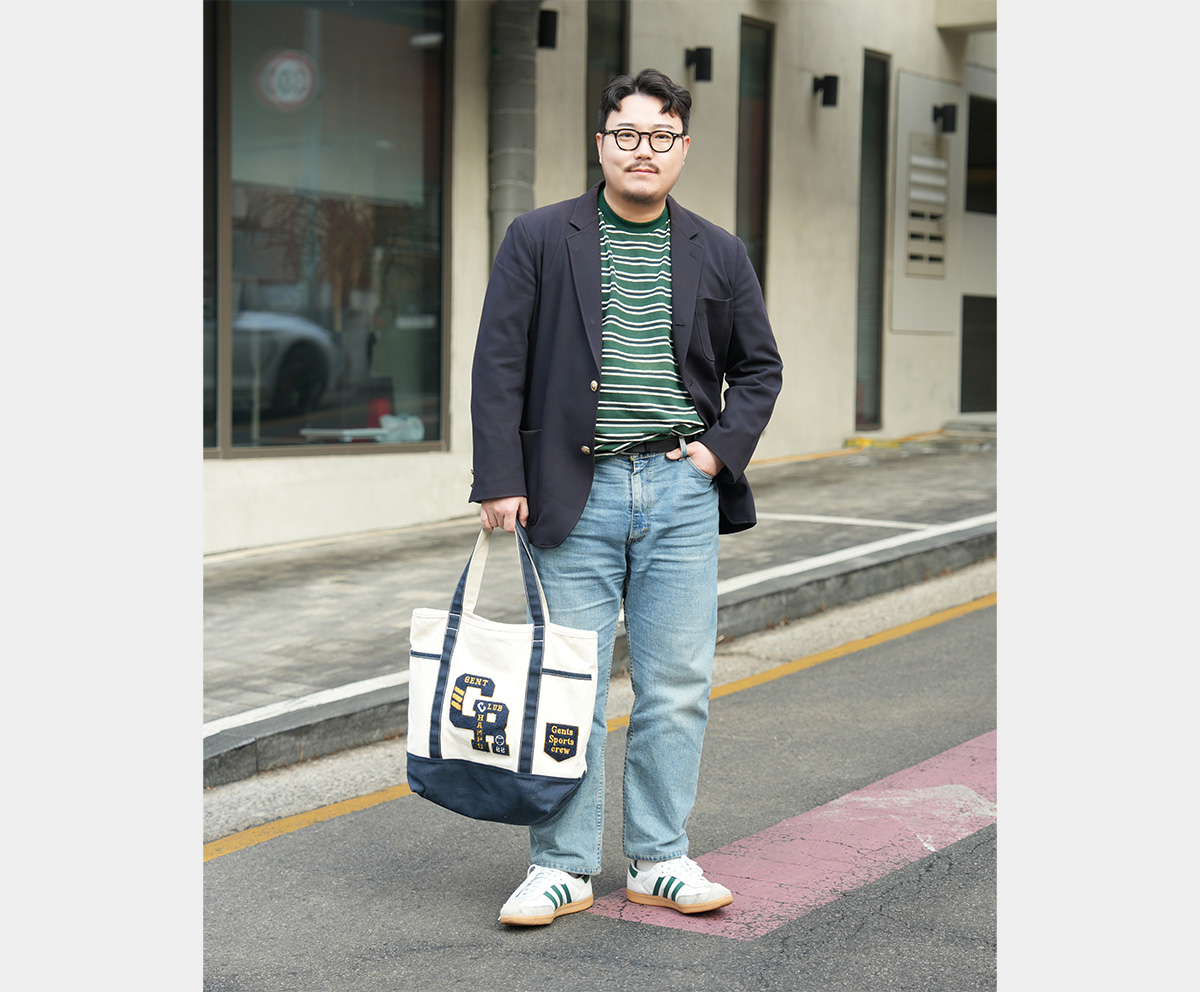 Full-body view of a man wearing a navy gold-button blazer with a striped T-shirt and light-wash denim.
