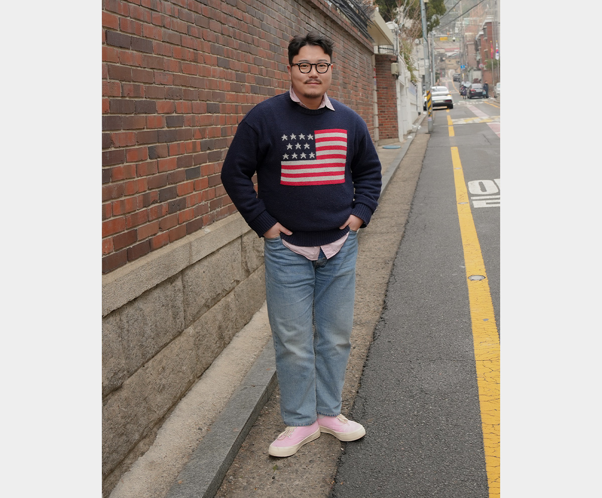 Full-body photo of a man standing in a navy knit with an American flag pattern and light-wash denim.