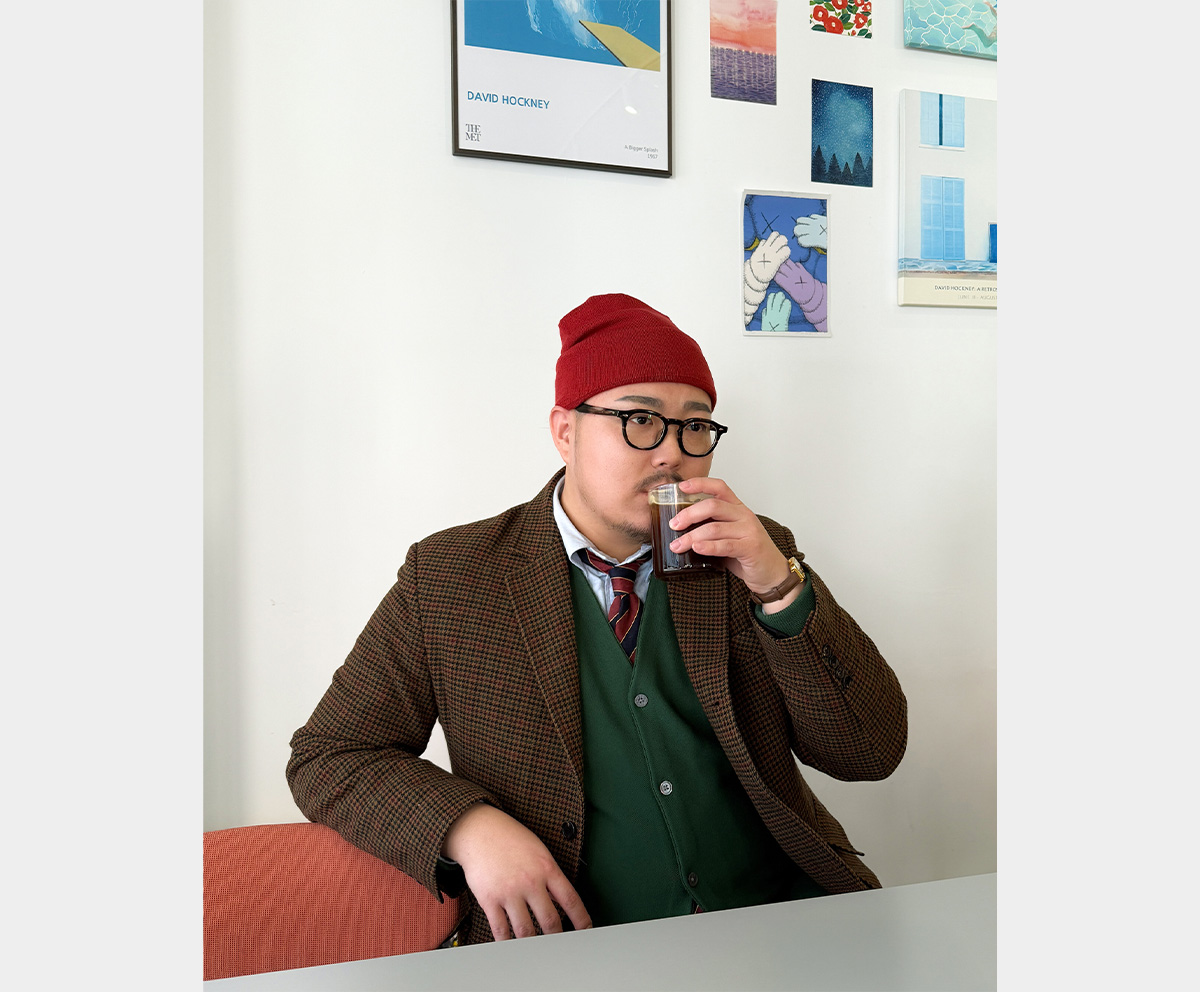 A man drinking coffee while wearing a houndstooth check jacket and a red beanie.
