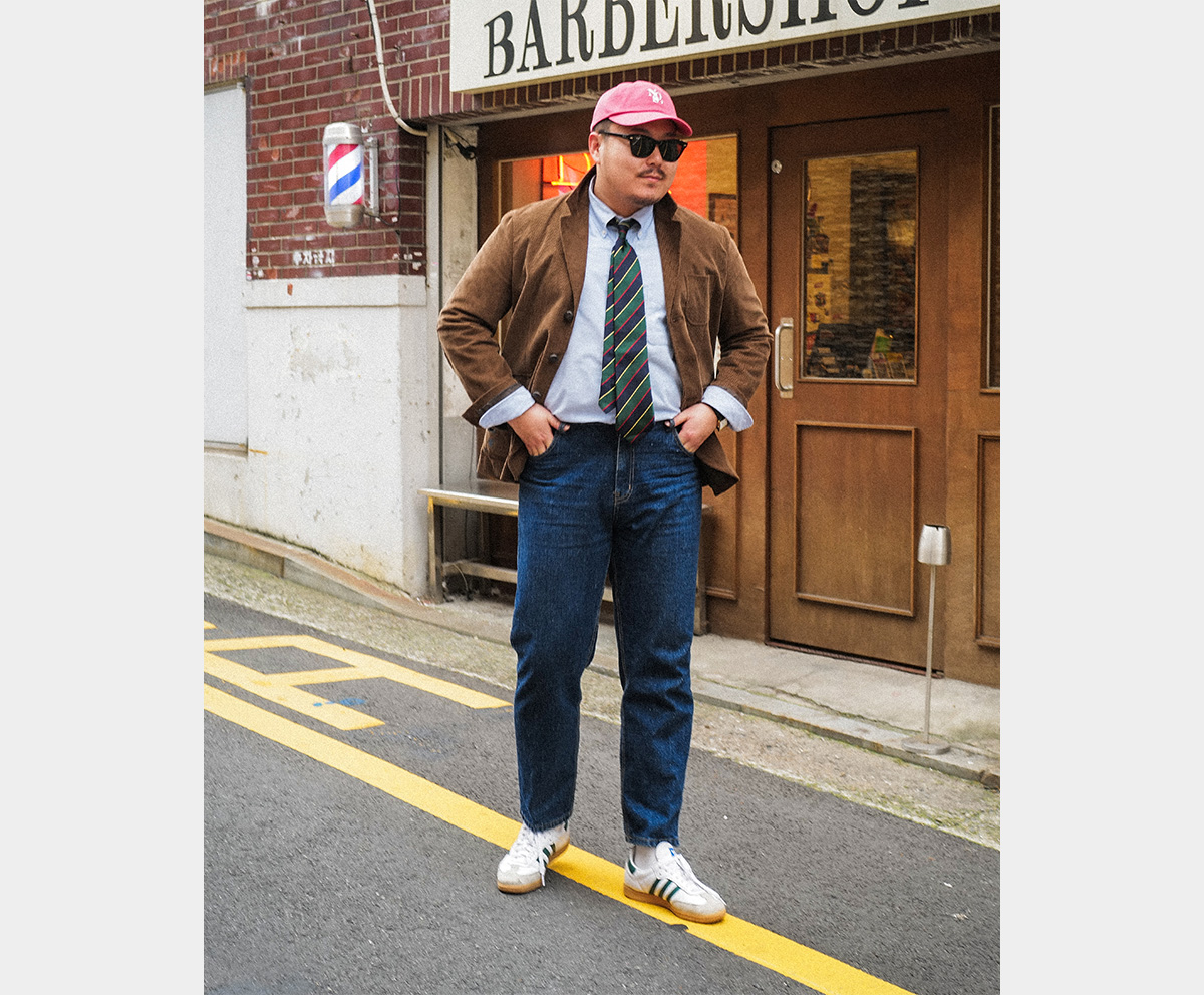 A man with his hands in his pockets, wearing a brown corduroy jacket and dark wash denim.