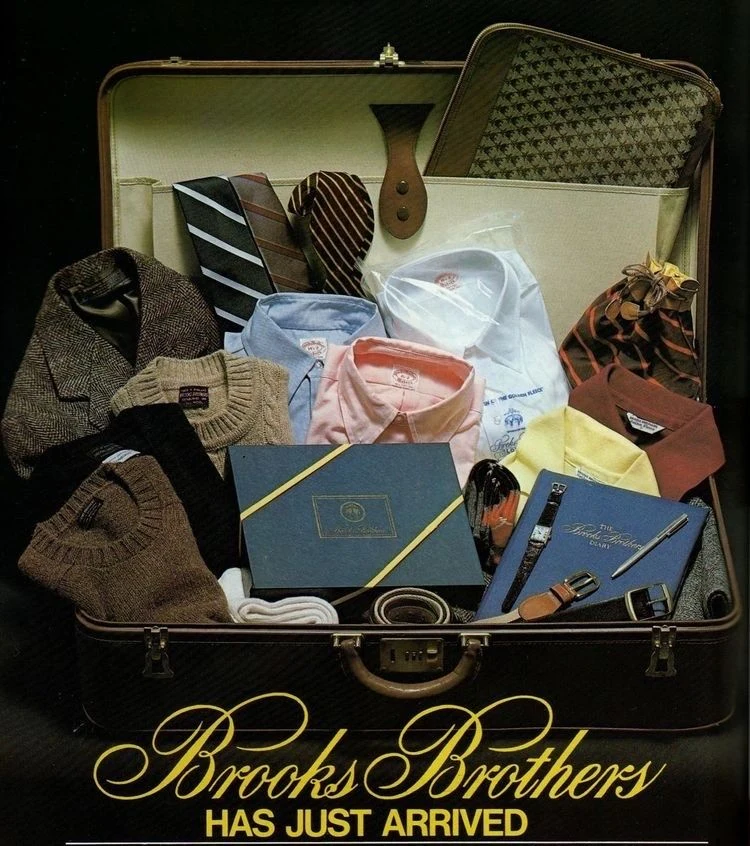 Brooks Brothers history: the iconic 'Has Just Arrived' vintage advertisement showing a suitcase packed with tweed jacket, Shetland sweaters, Original Polo button-down shirts in white, blue, pink and yellow, repp ties, and the Brooks Brothers diary — the essential Brooks Brothers wardrobe