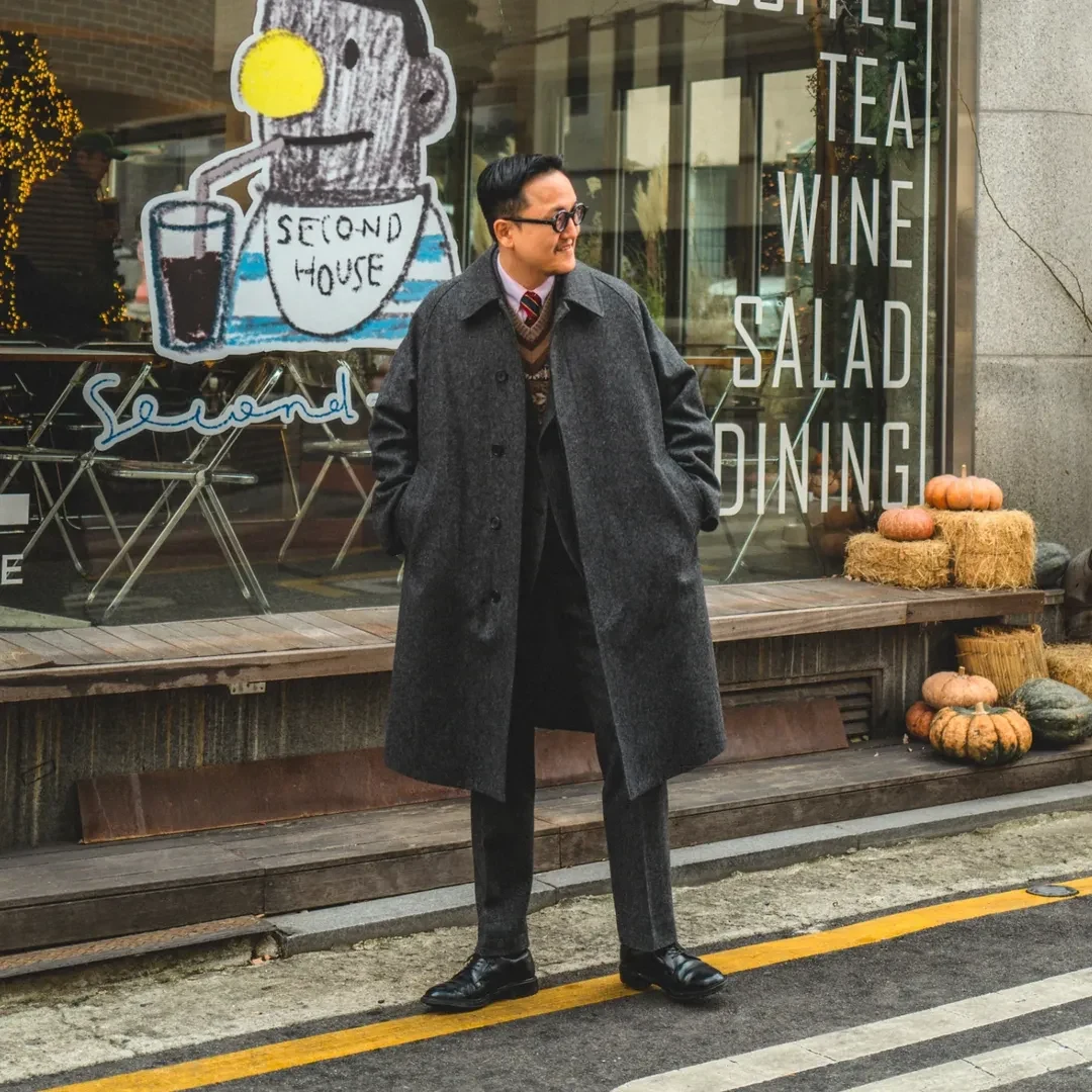 How to wear a Fair Isle vest under a balmacaan coat: charcoal grey balmacaan coat over a charcoal grey suit with Fair Isle vest underneath, OCBD, regimental rep tie, and black plain-toe derbies