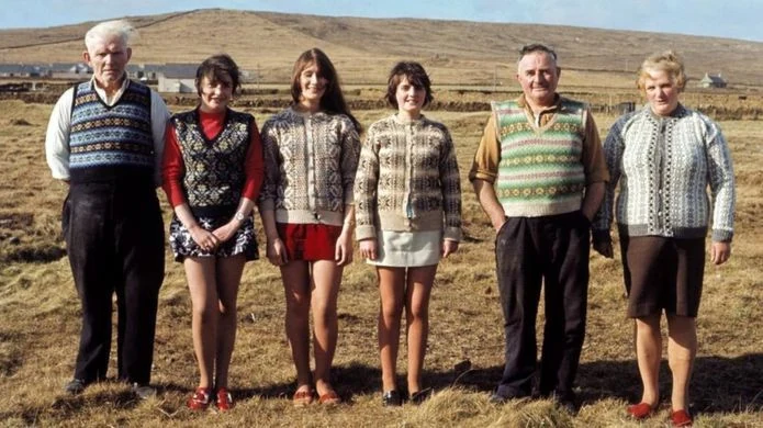 How to wear a Fair Isle vest origin: vintage color photograph of a six-person Shetland family on Fair Isle island, each wearing a different traditional hand-knit Fair Isle pattern — vests, cardigans, and pullovers — standing together in a coastal field