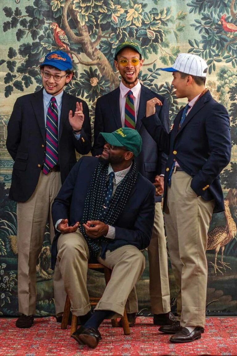 Four young men in modern American Ivy editorial — navy gold-button blazers with patch pockets, OCBDs, regimental rep ties, khaki chinos, ballcaps, penny loafers — natural shoulder sack-cut tradition photographed on a tapestry backdrop
