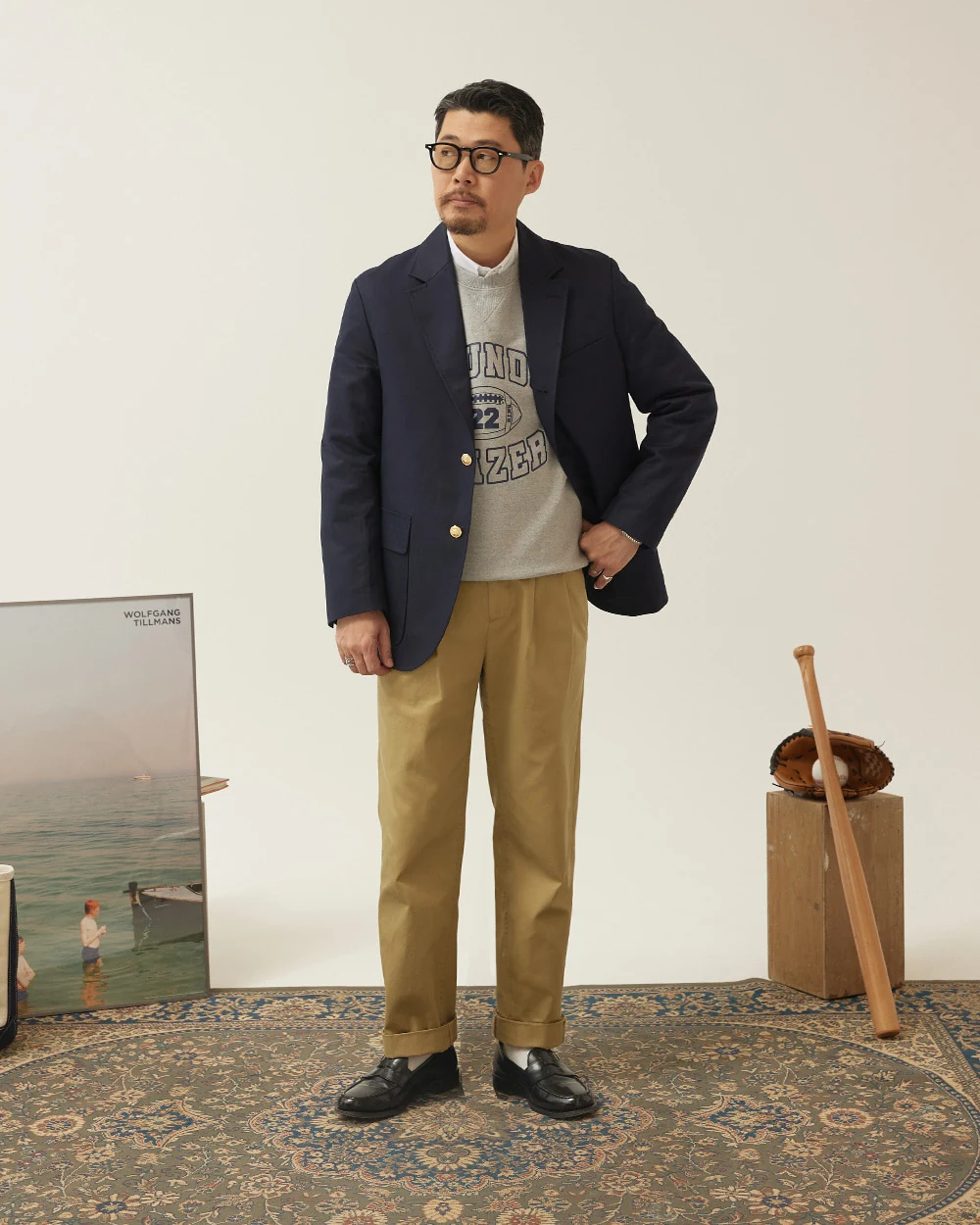 Navy gold-button blazer outfit — single-breasted blazer, grey sweatshirt layered over white OCBD, khaki chinos, plaque belt, black penny loafers, modern casual layered configuration