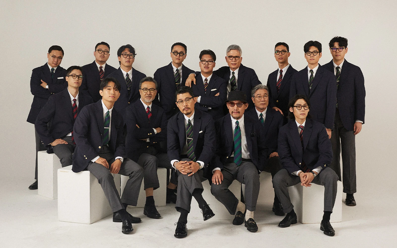 Renacts Legacy Suits campaign Configuration B group — same sixteen customers in navy gold-button double-breasted blazers with grey trousers, eight seated and eight standing
