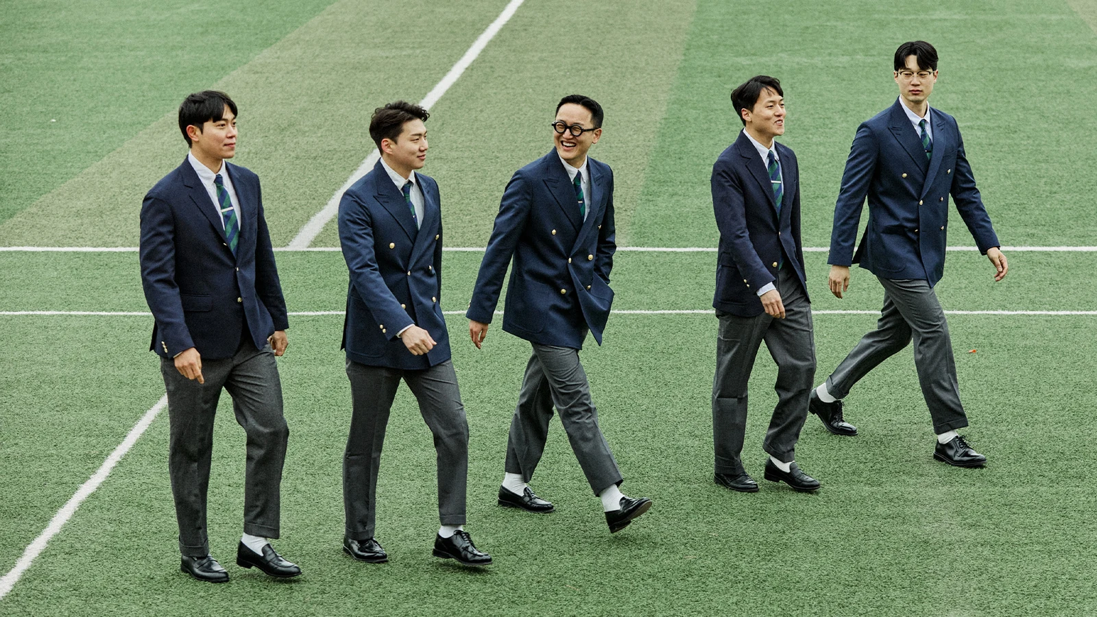 Five Renacts customers walking in double-breasted navy gold-button blazer outfits with grey wool trousers, photographed outdoor on a football field