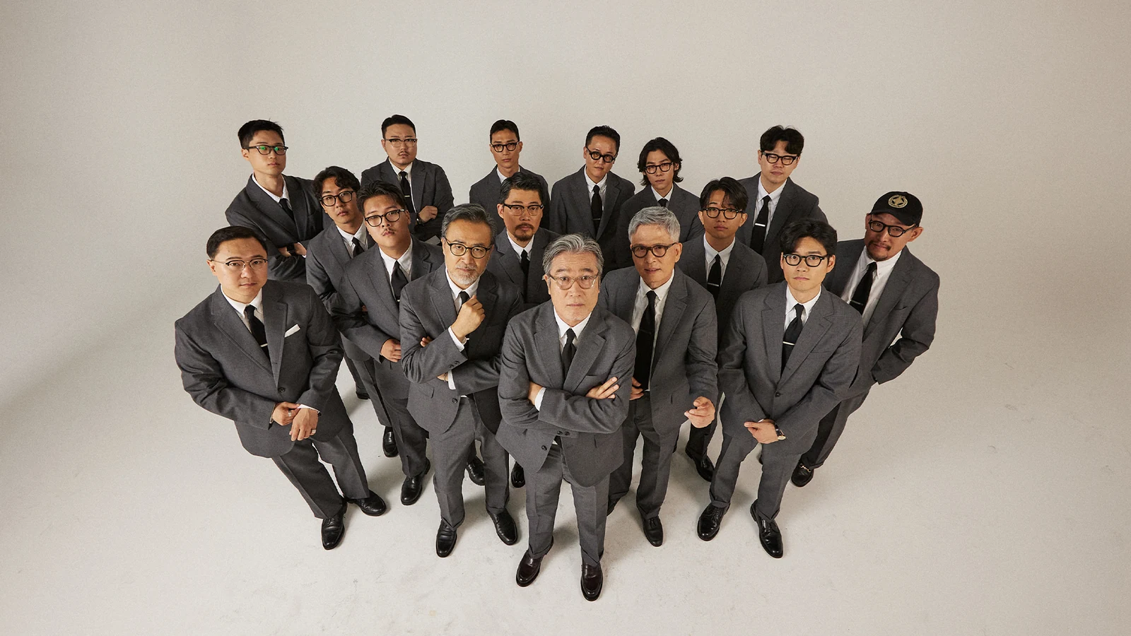 Renacts Legacy Suits 2023 Fall campaign group portrait: sixteen Gentlemens Club members aged 20s to 60s photographed from above, each in a grey wool sack suit, white oxford button-down, regimental rep tie, and black leather shoes