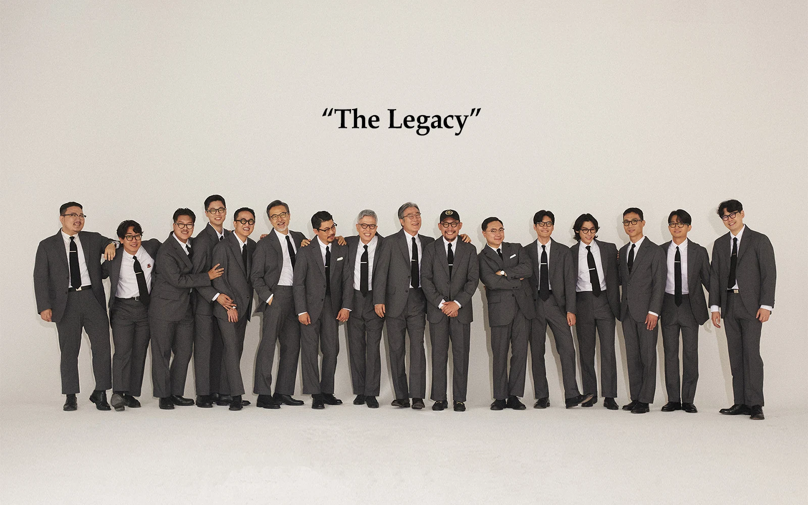Renacts Legacy Suits 2023 campaign 'The Legacy' titled hero portrait — sixteen Gentlemens Club members aged 20s to 60s, standing in a single horizontal line in grey wool sack suits, white OCBDs, and black silk knit ties, with the words 'The Legacy' set above them