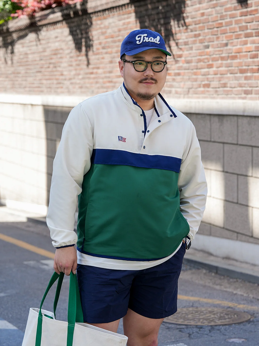 Renacts Trad cap portrait — navy six-panel cap with cursive Trad embroidery, tortoise round eyewear, and bicolor anorak placket