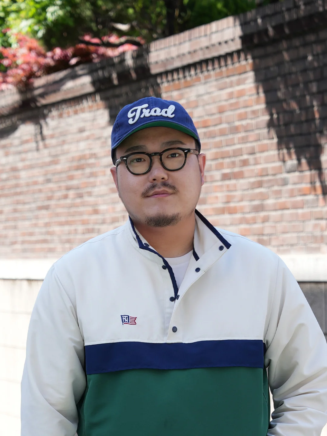 Renacts Trad cap close portrait — cursive Trad script embroidery, round tortoise frames, and Renacts bicolor anorak with chest placket