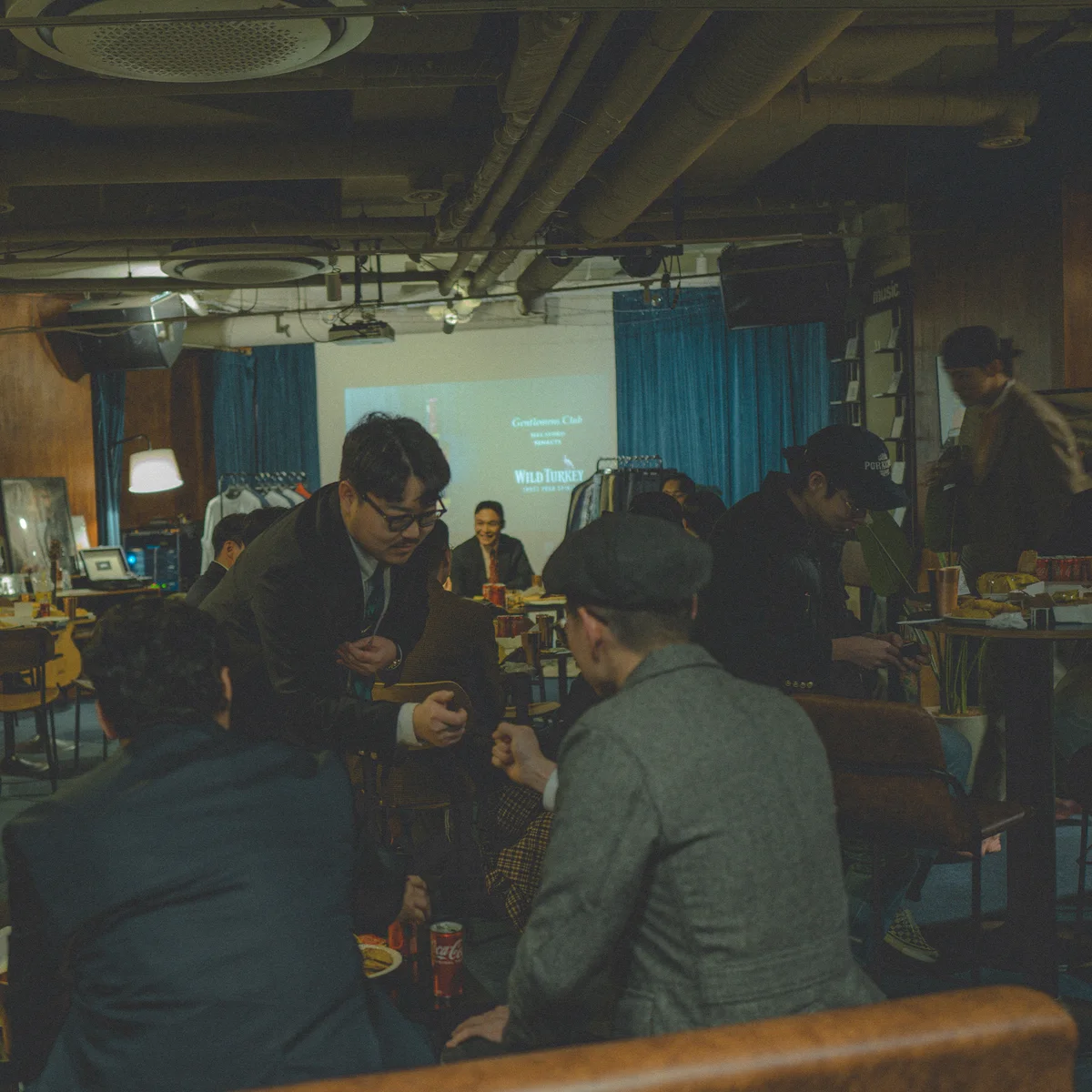 Gentlemen's House venue wide shot — Gentlemens Club Wild Turkey projection on the back wall, members gathered for the sixth meeting