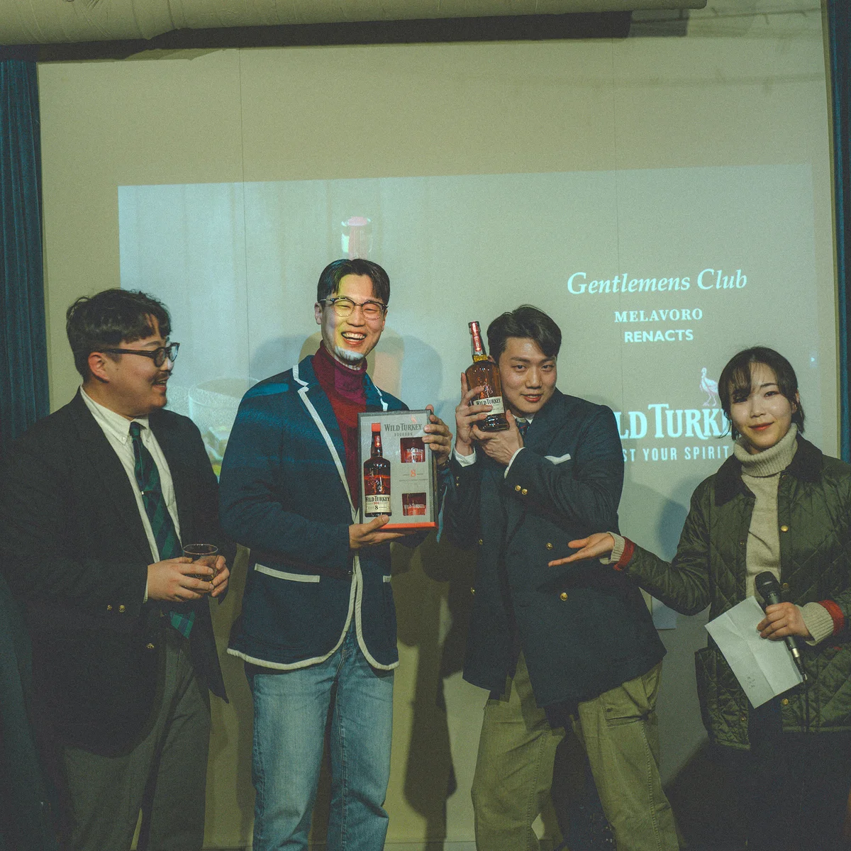 A curator from Wild Turkey holding the eight-year box on stage, with the night's host slide for Gentlemens Club, Melavoro, and Renacts projected behind