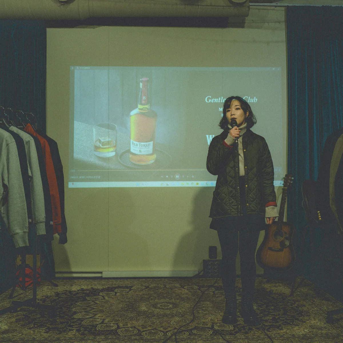 Melavoro curator at the microphone — Wild Turkey label projected on the back wall, vintage rail and an acoustic guitar in frame