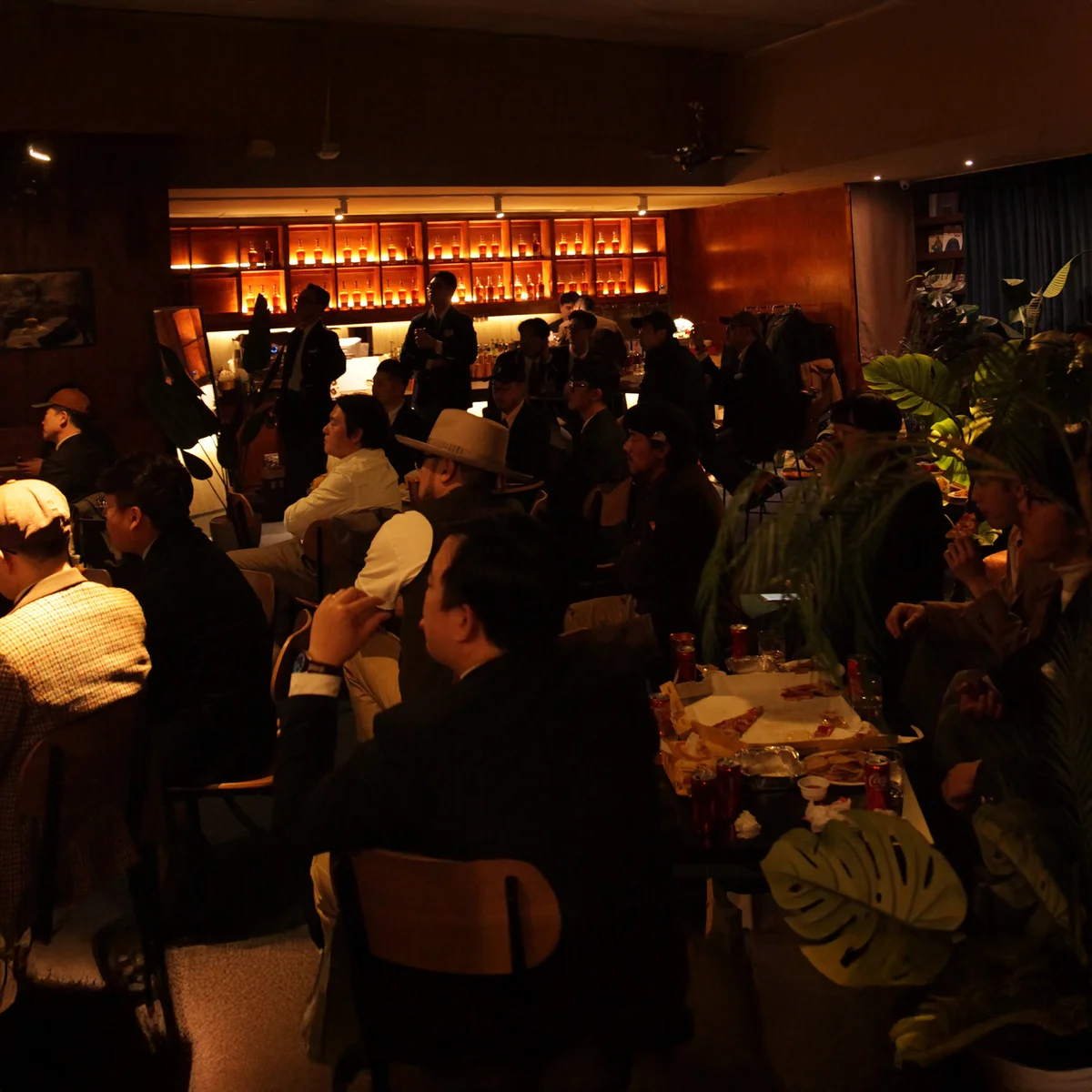 Wide shot of Gentlemen's Club Vol. 6 evening — fifty members at the long table and bar, lit warmly