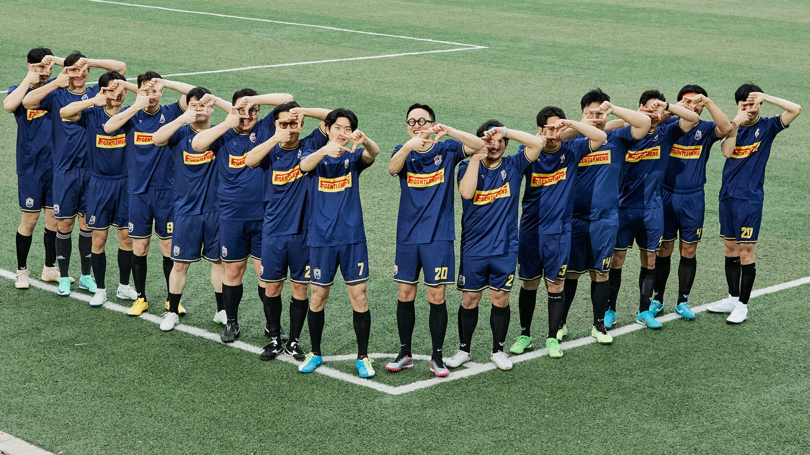 Gentlemens Football Club jersey salute lineup: sixteen members in navy GENTLEMENS football kits hands raised to forehead in salute formation post-match at Guri Wangsuk Sports Park centre line