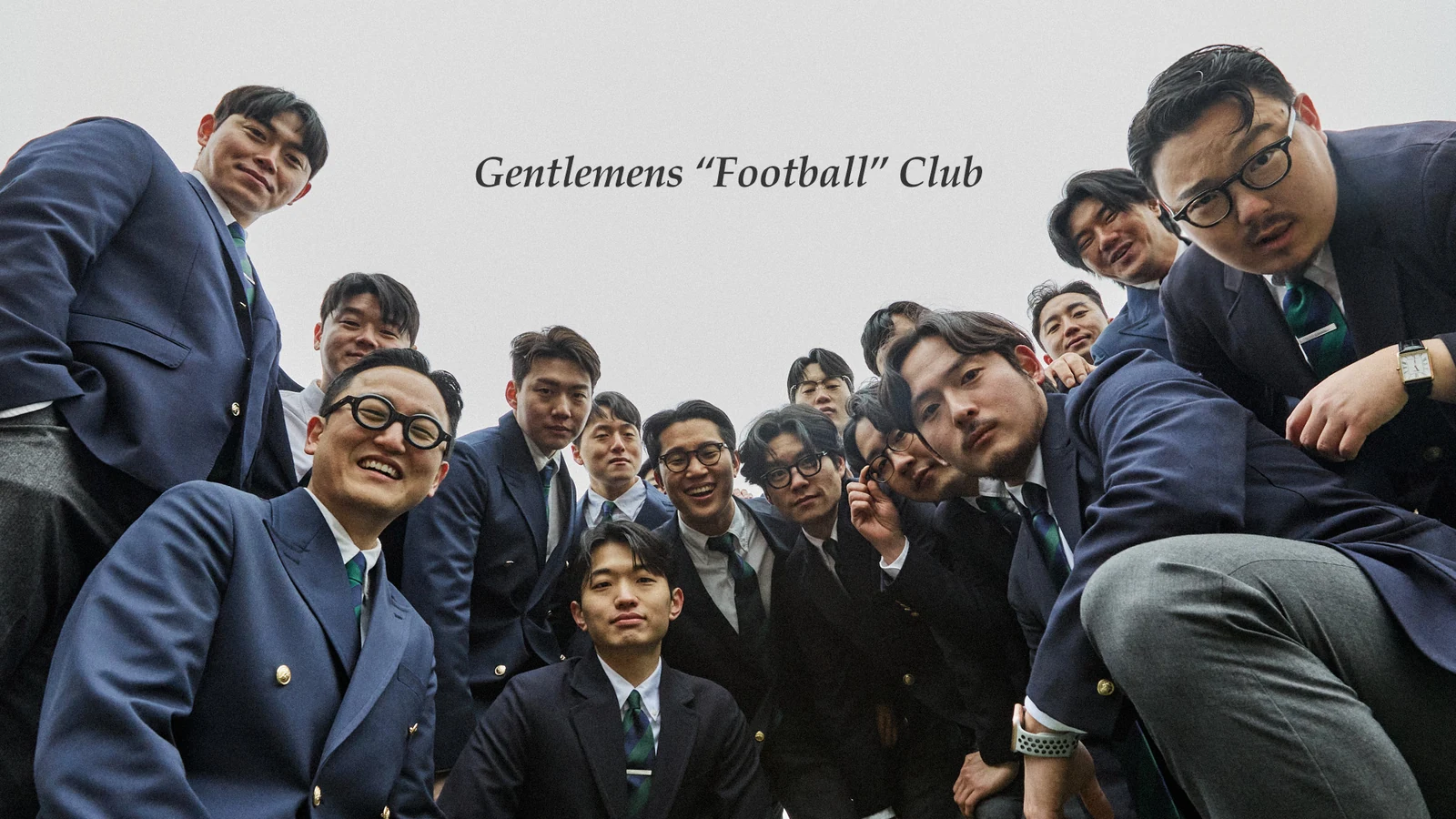 Gentlemens Football Club team huddle from below: sixteen members in navy blazers looking down at camera Gentlemens Football Club title text sky background closing campaign frame