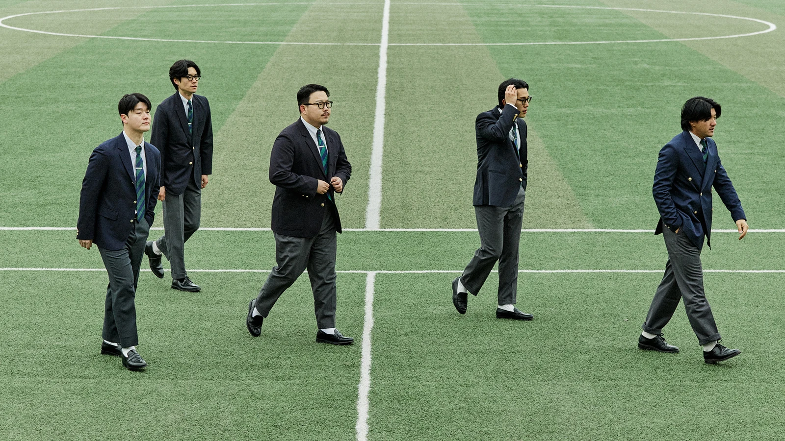 Gentlemens Football Club walking field diagonal: five members in navy blazers and grey trousers walking across the centre line of Guri Wangsuk Sports Park before the match