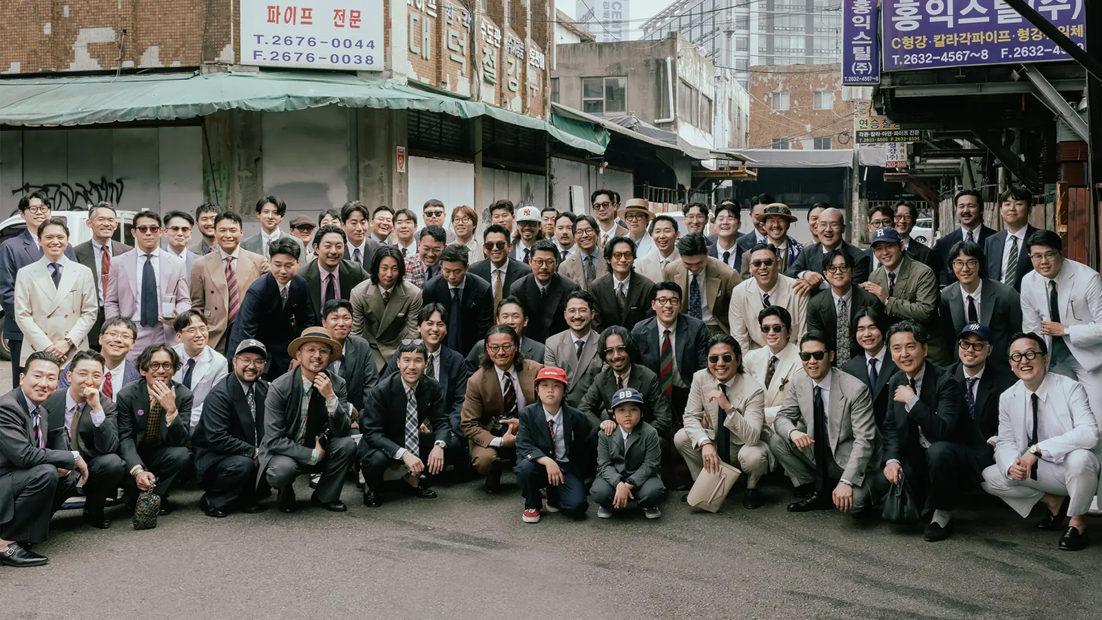 Around eighty people in tailored trad and Ivy outfits, posed in rows across a street of old steel-supply shops in central Seoul, two children in suits at the front.