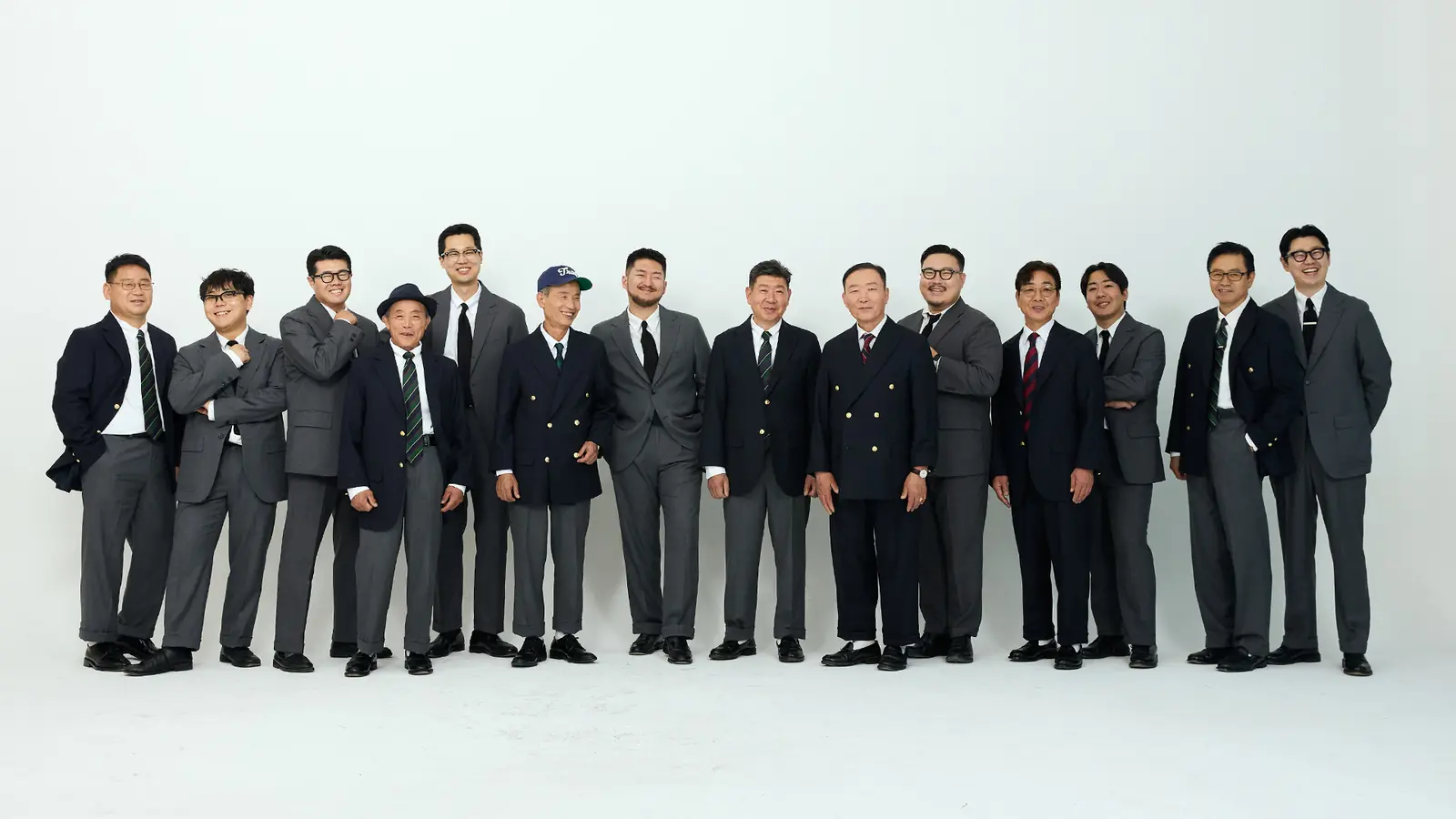 A line of about fifteen men against a white studio wall, some in navy blazers with gilt buttons, some in grey flannel suits, all in white shirts with striped or knitted ties, ranging from men in their twenties to men in their seventies.