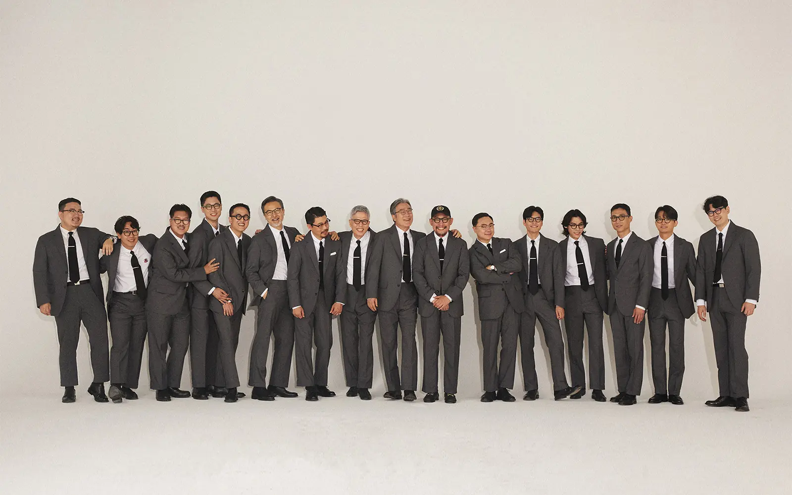 A long line of men in grey flannel sack suits with white shirts and black knit ties, leaning on one another against a plain studio wall.