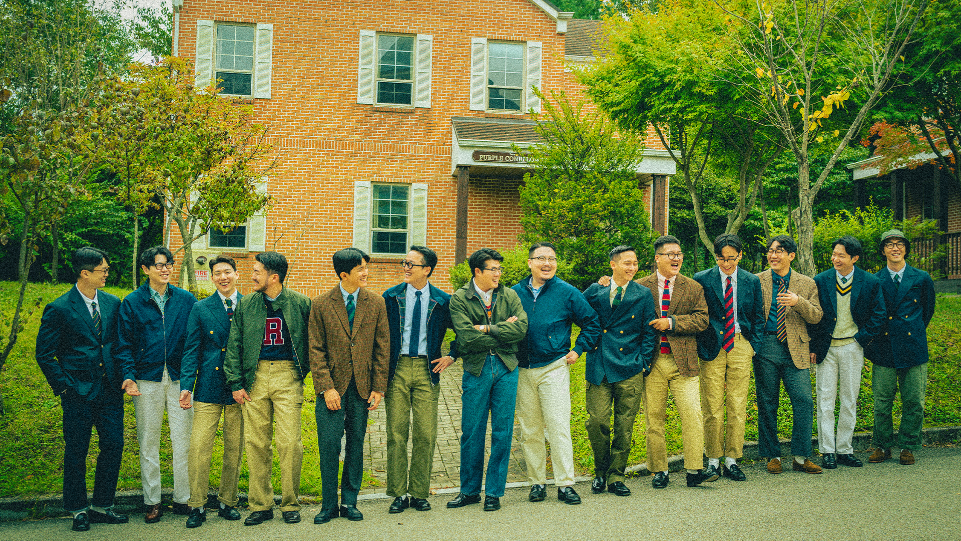 A line of men in navy blazers and tweed jackets, button-down shirts, ties and khaki chinos in front of a brick campus building, Renacts Take Ivy campaign, Korea 2023