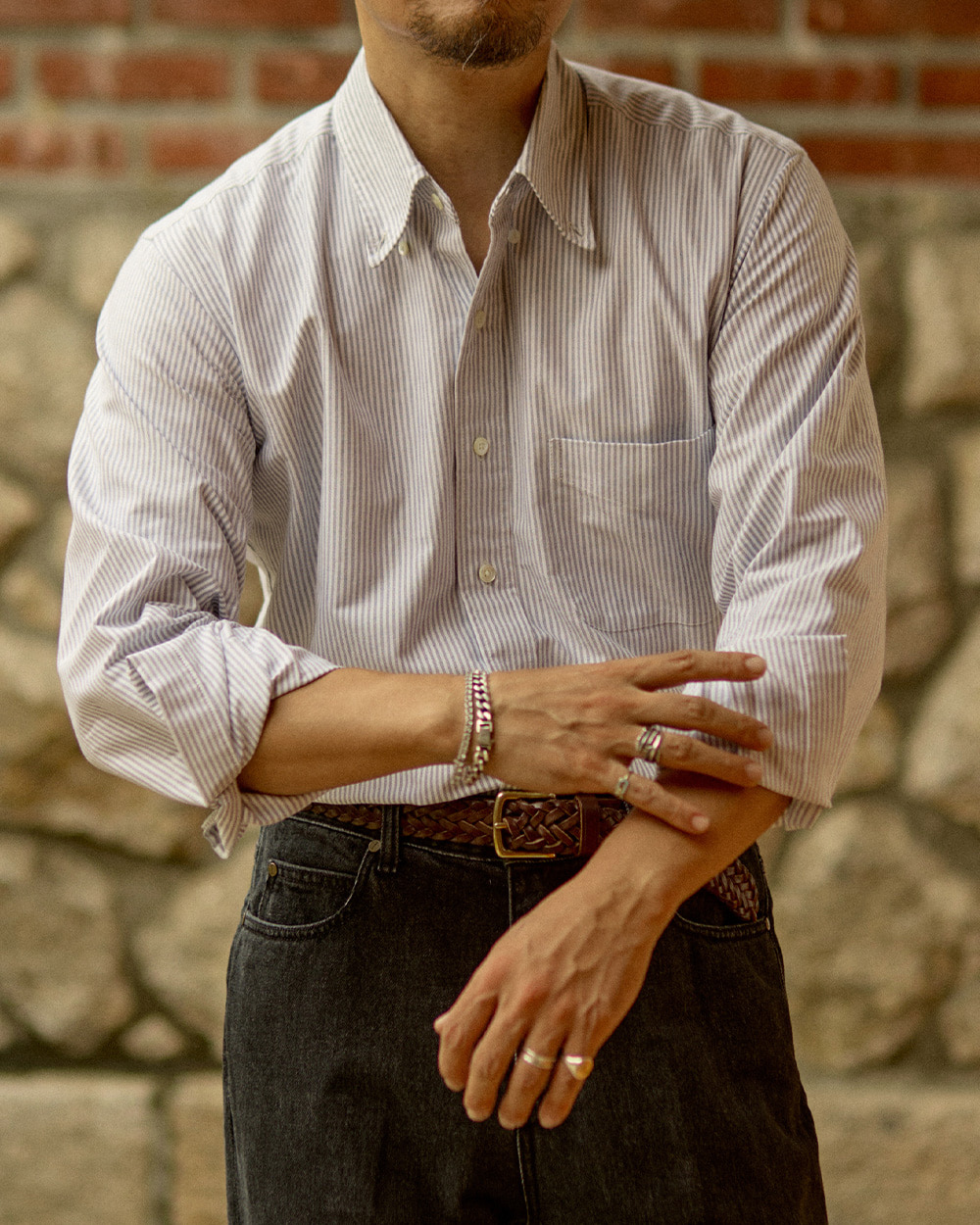 Cream striped oxford button-down shirt worn open at the collar with sleeves rolled, in front of a stone wall in Seoul