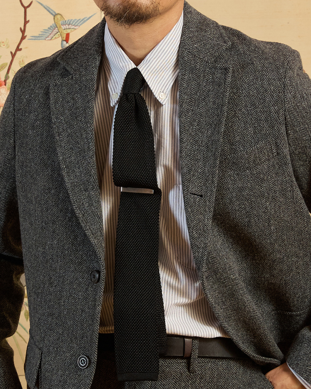 Striped oxford button-down shirt and black knit tie worn under a grey tweed jacket, collar roll visible above the lapels