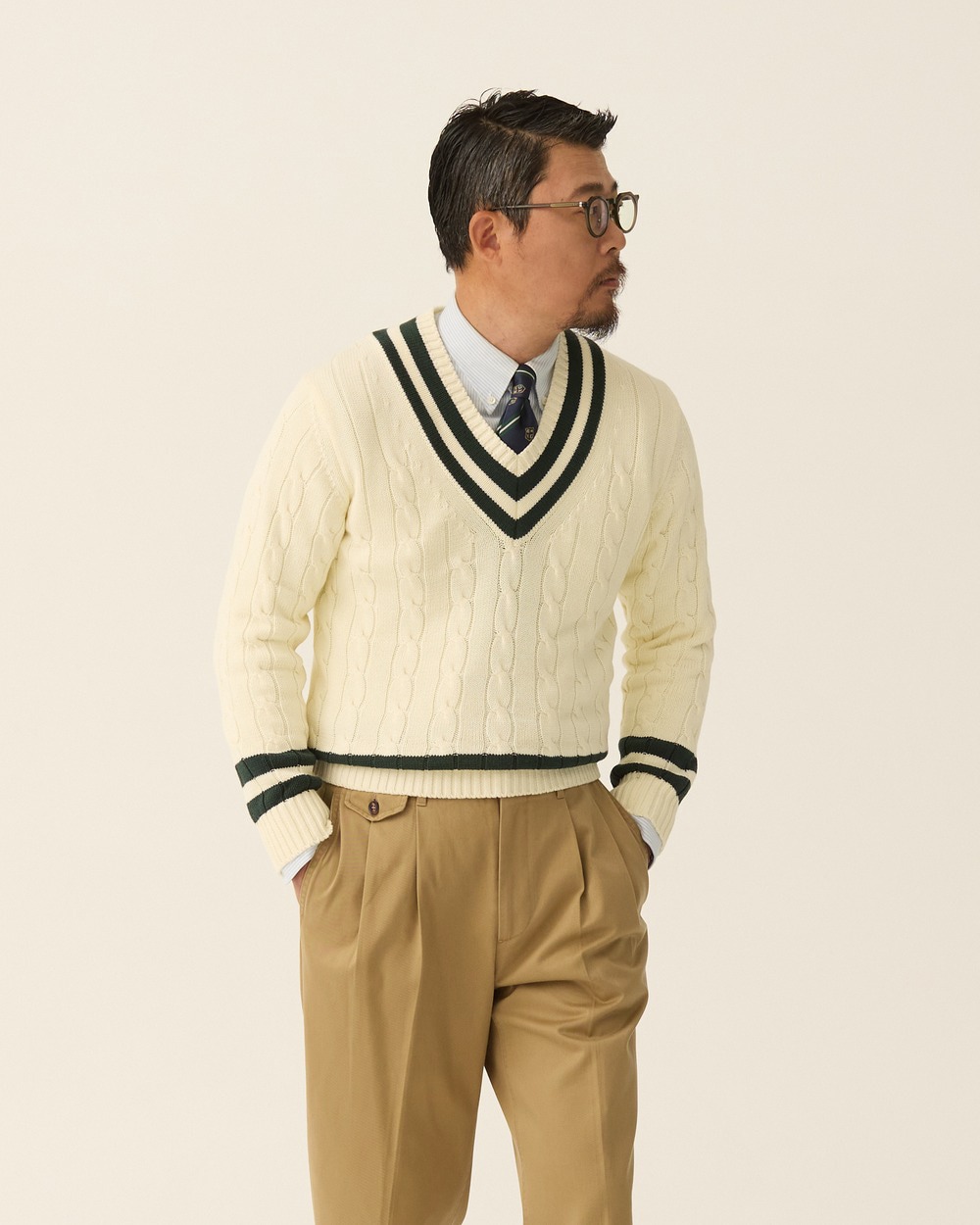 Close view of an ivory cricket sweater with green and navy trim worn over a shirt and striped tie with pleated khaki chinos