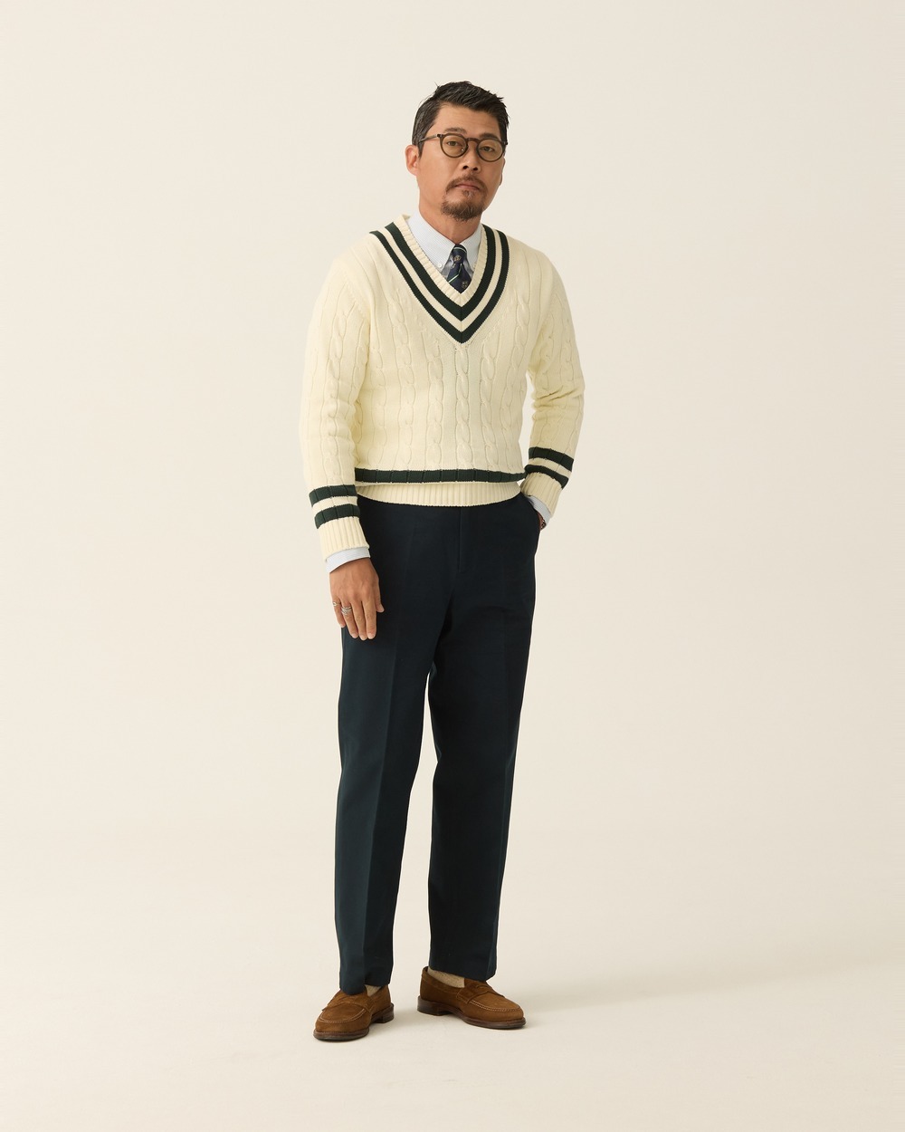 Ivory cable-knit cricket sweater with green and navy trim at the V-neck, cuffs and hem, worn over a shirt and striped tie with navy trousers and brown suede loafers