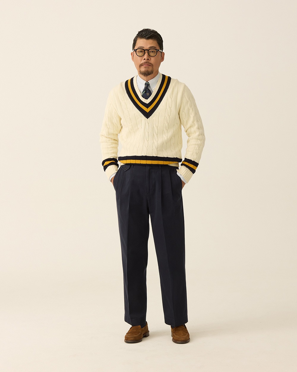 Ivory cricket sweater with navy and gold trim worn over a shirt and striped tie, with navy trousers and brown suede loafers