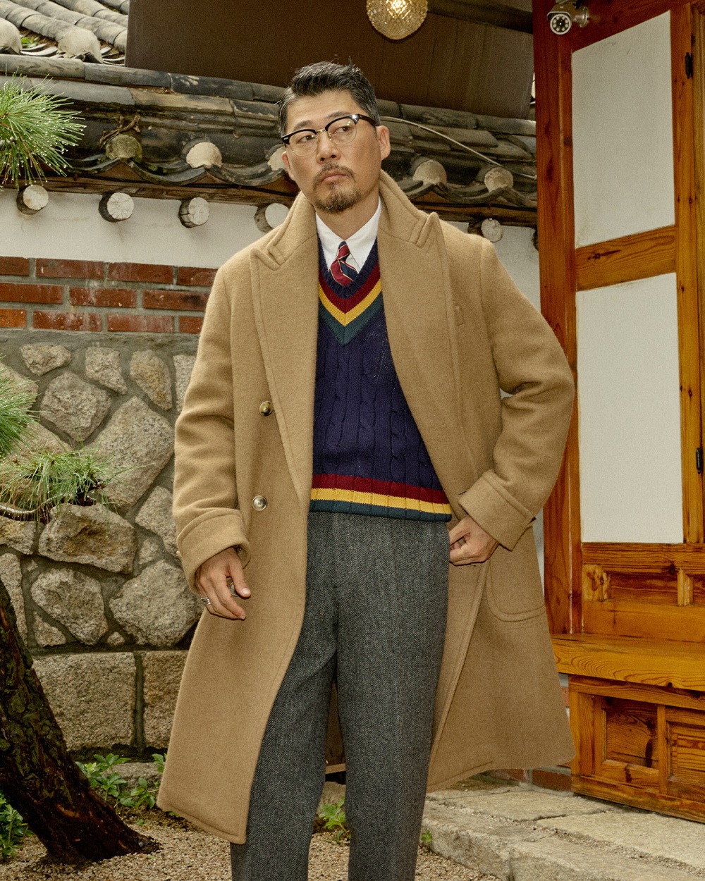 Camel overcoat worn open over a navy cricket sweater with a shirt and tie and grey herringbone trousers, in front of a hanok wall