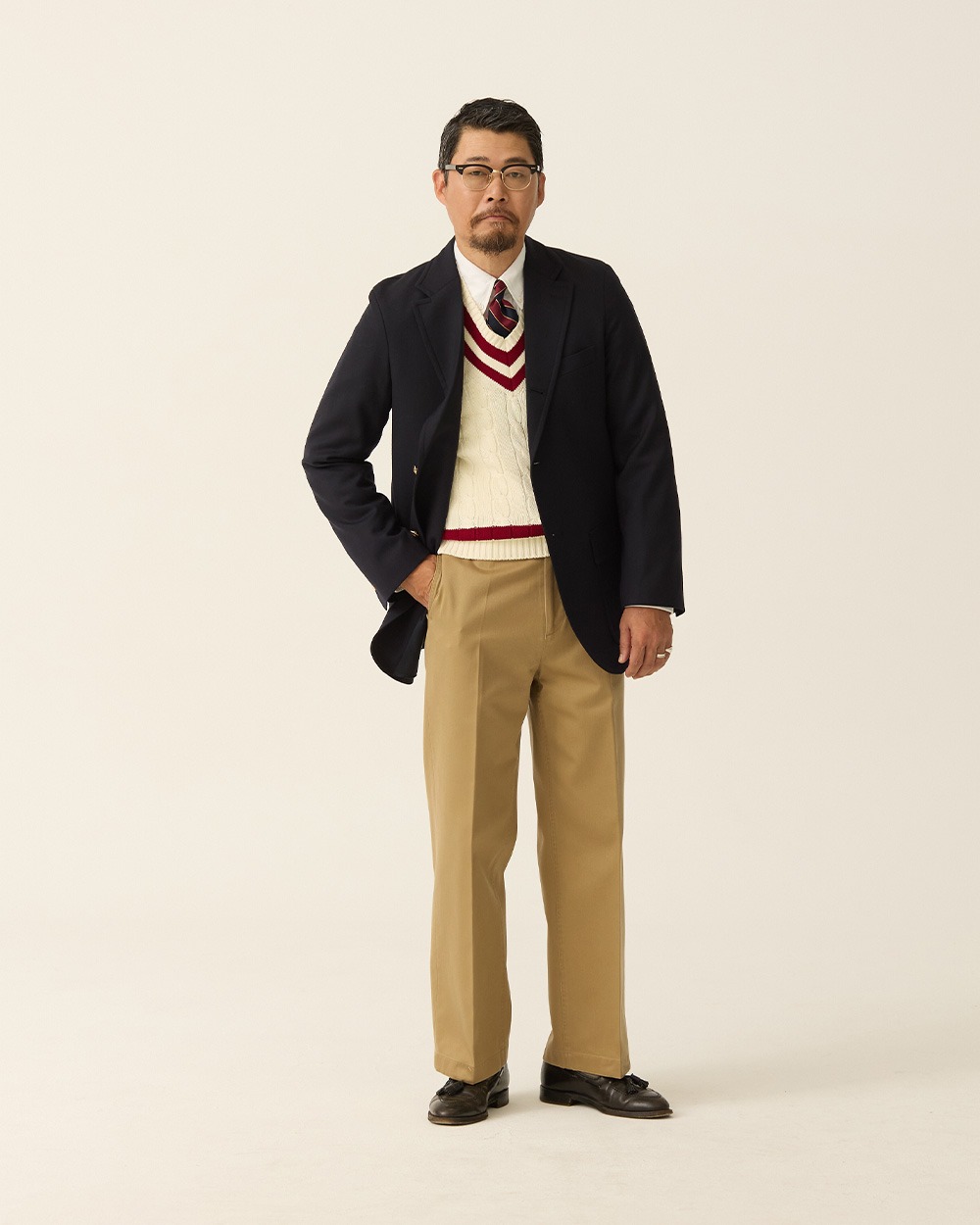 Navy blazer worn open over an ivory cricket sweater with red trim, a shirt and tie, khaki chinos and dark loafers