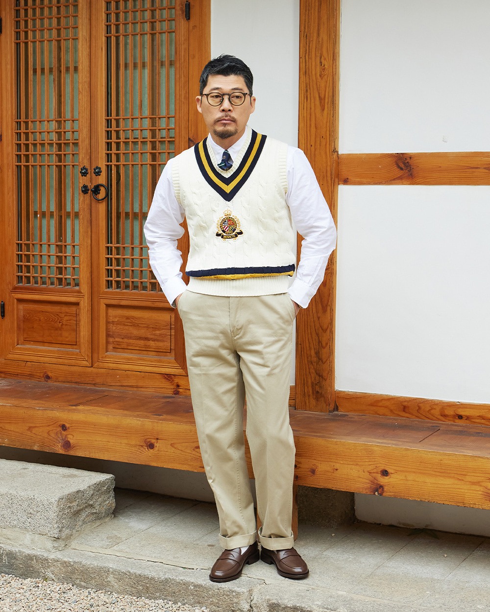 Ivory crest cricket vest worn over a white shirt and tie with khaki chinos, in front of a hanok doorway