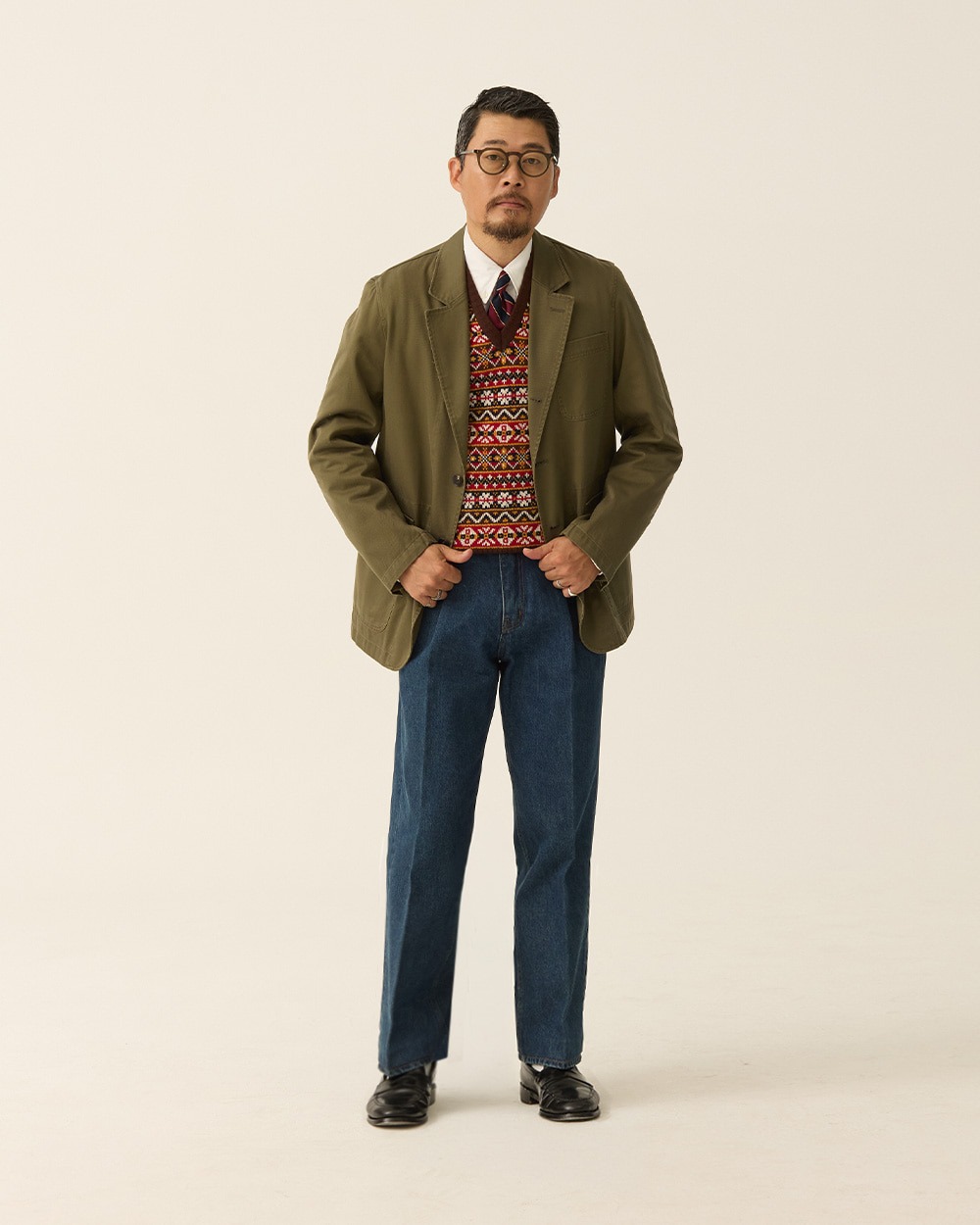 Olive jacket worn open over a Brown Multi Fair Isle vest, white shirt and tie, with straight indigo jeans and black loafers