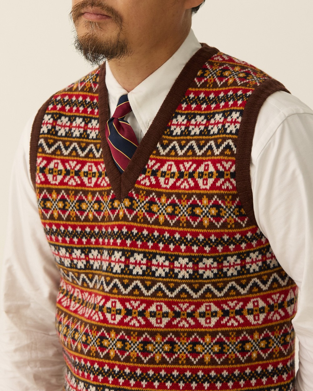 Close-up of a Fair Isle vest with red, white, gold and green pattern bands inside dark brown ribbing, worn over a white shirt with a striped repp tie