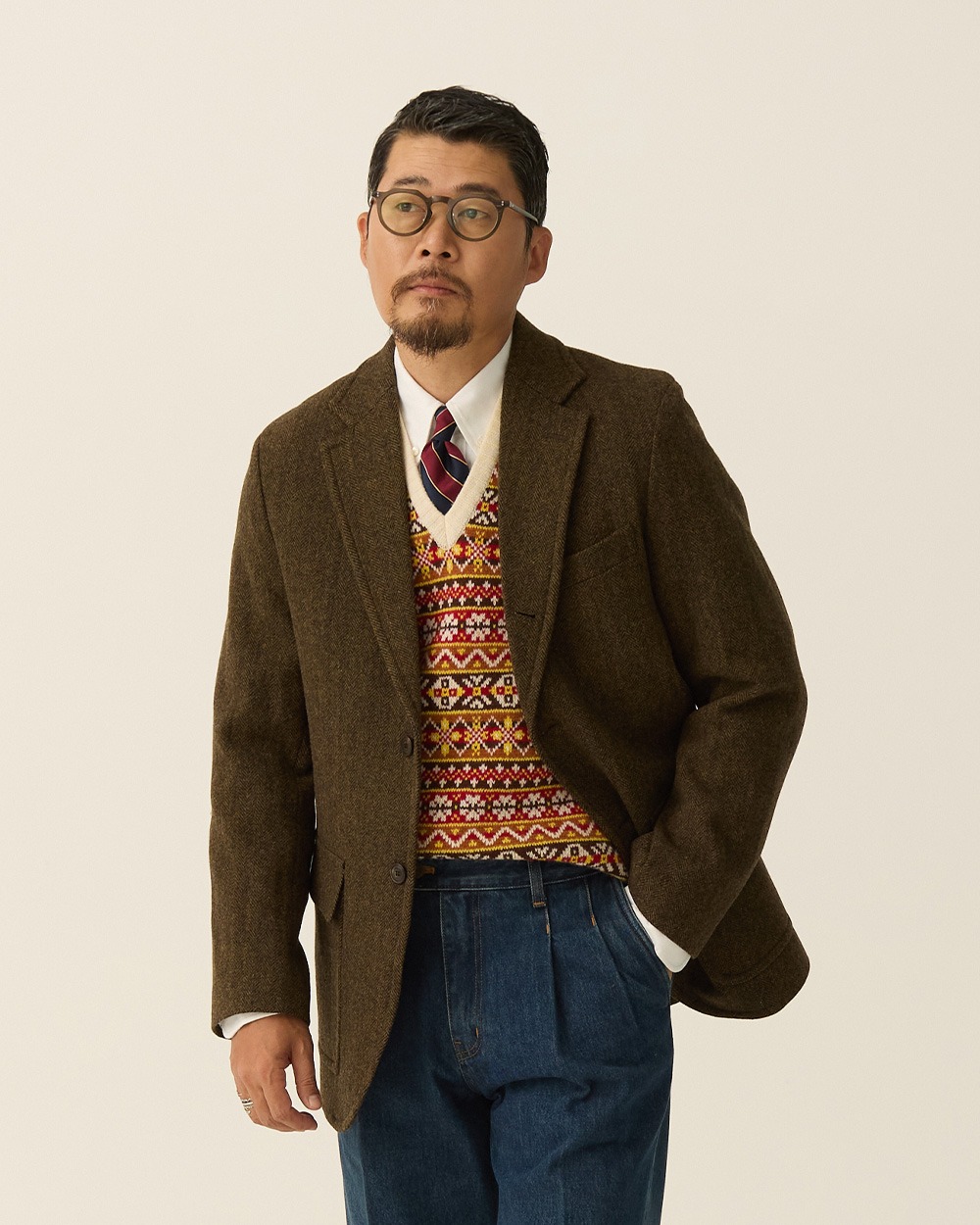 Brown tweed jacket worn open over an Ivory Multi Fair Isle vest, white shirt and striped tie, with indigo jeans