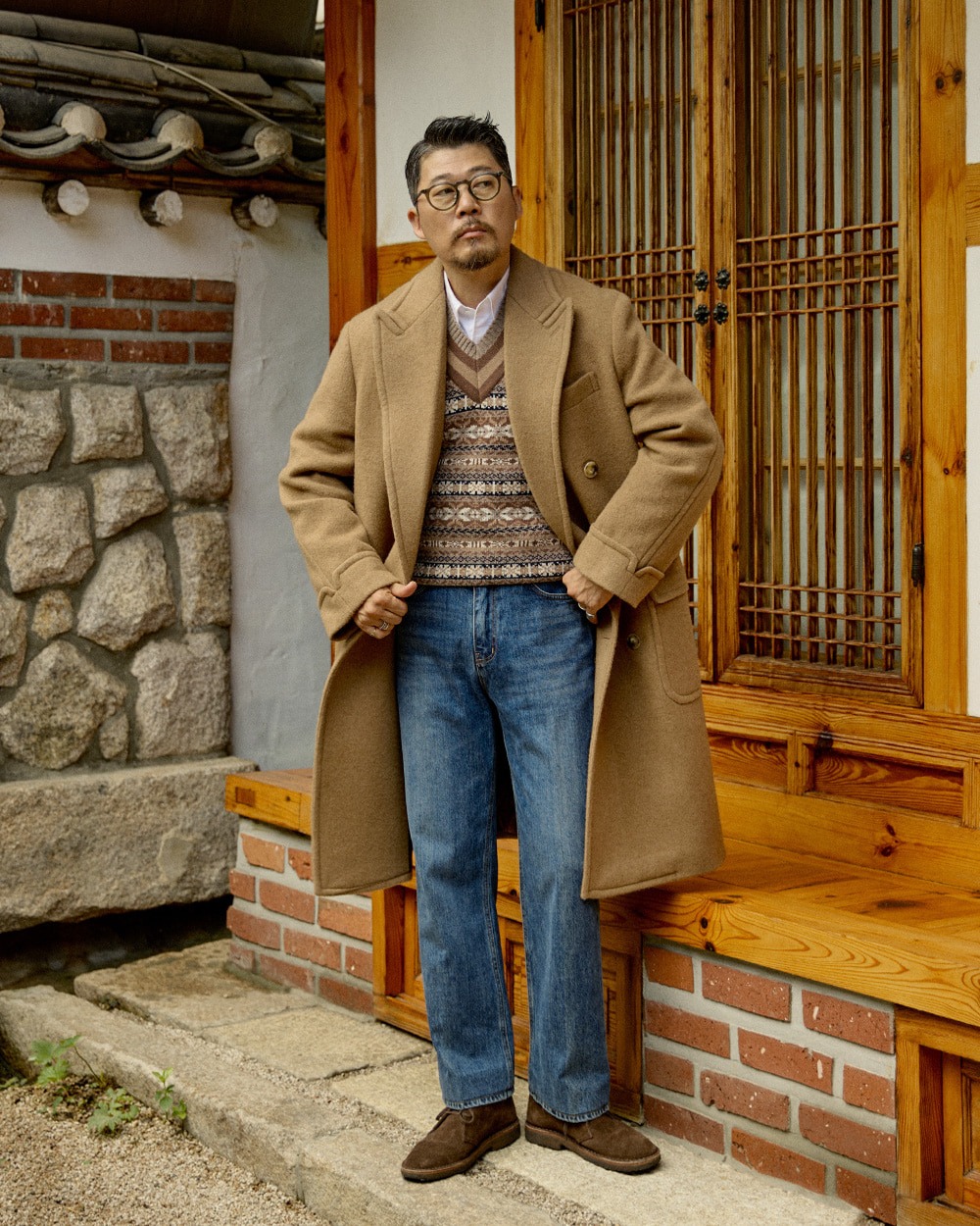 Camel overcoat worn open over the original taupe Fair Isle vest and a white shirt, with straight indigo jeans and brown suede shoes