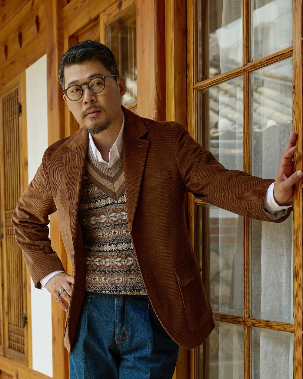 Original taupe Fair Isle vest over an open-collar white shirt under a brown corduroy jacket with indigo jeans, beside a hanok window