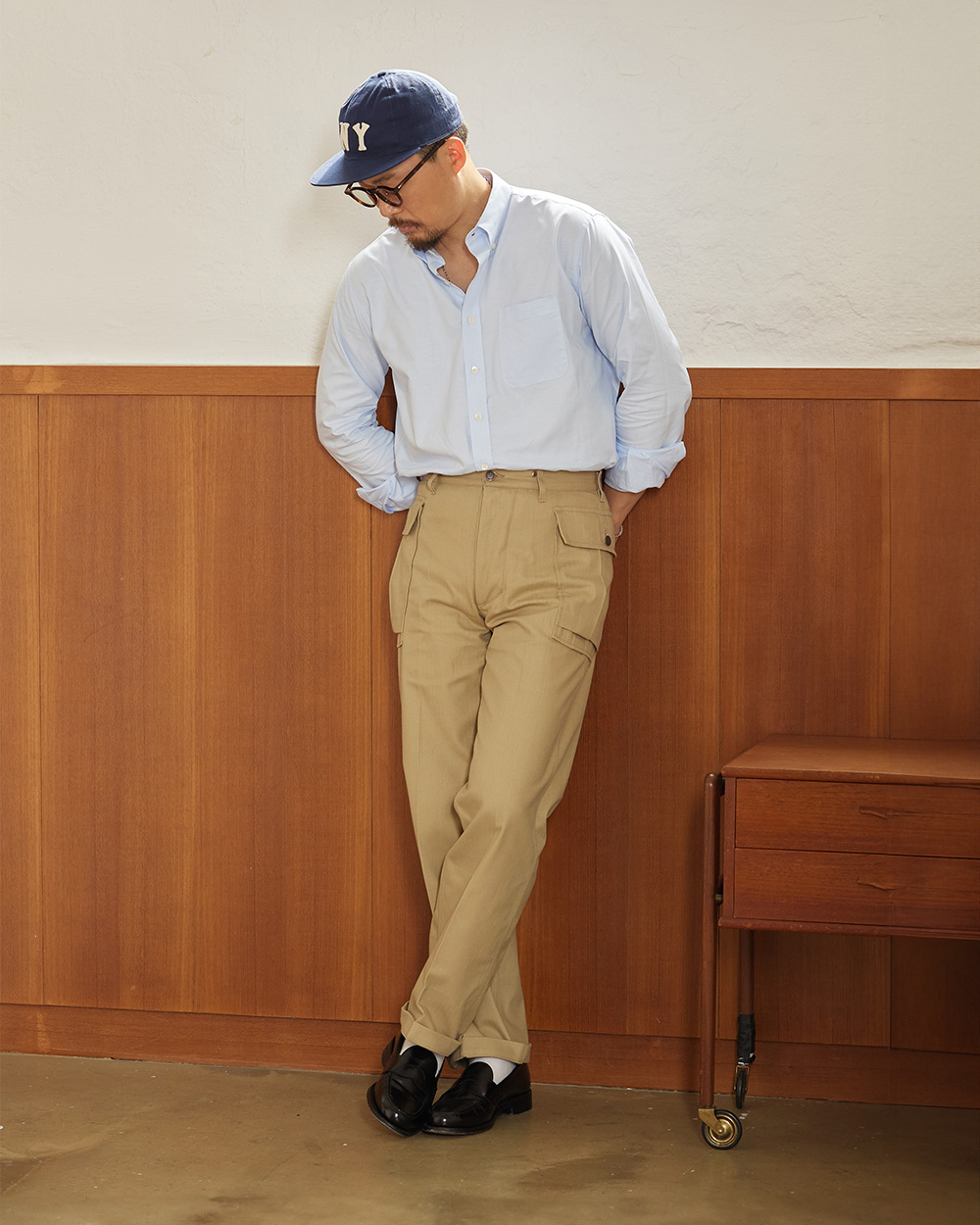 Light blue oxford button-down shirt tucked into tan chinos with a navy ball cap and penny loafers, leaning against a wood-paneled wall in Seoul
