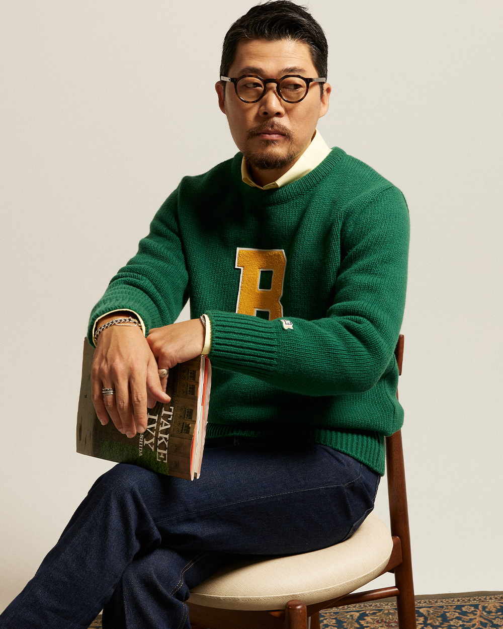 modern ivy style: green boat-neck letterman sweater with a chenille varsity letter worn over a collared shirt with indigo denim, holding the Take Ivy photobook