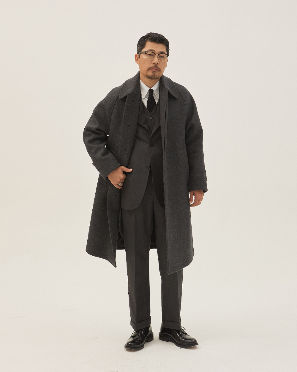 modern ivy winter layering: grey herringbone three-piece with cuffed trousers under a charcoal wool balmacaan coat with white shirt and black tie