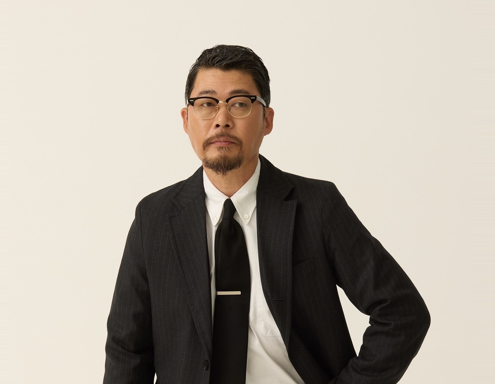 Upper body of a man in a charcoal pinstripe sack jacket with a white button-down shirt, a dark tie and a tie bar, one hand on his hip