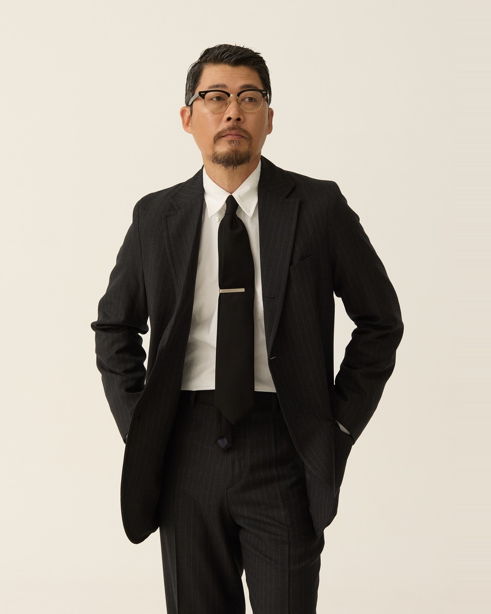 Man wearing a charcoal pinstripe sack jacket with a white shirt, a dark tie and a tie bar, hands in his trouser pockets, the jacket hanging straight from the chest
