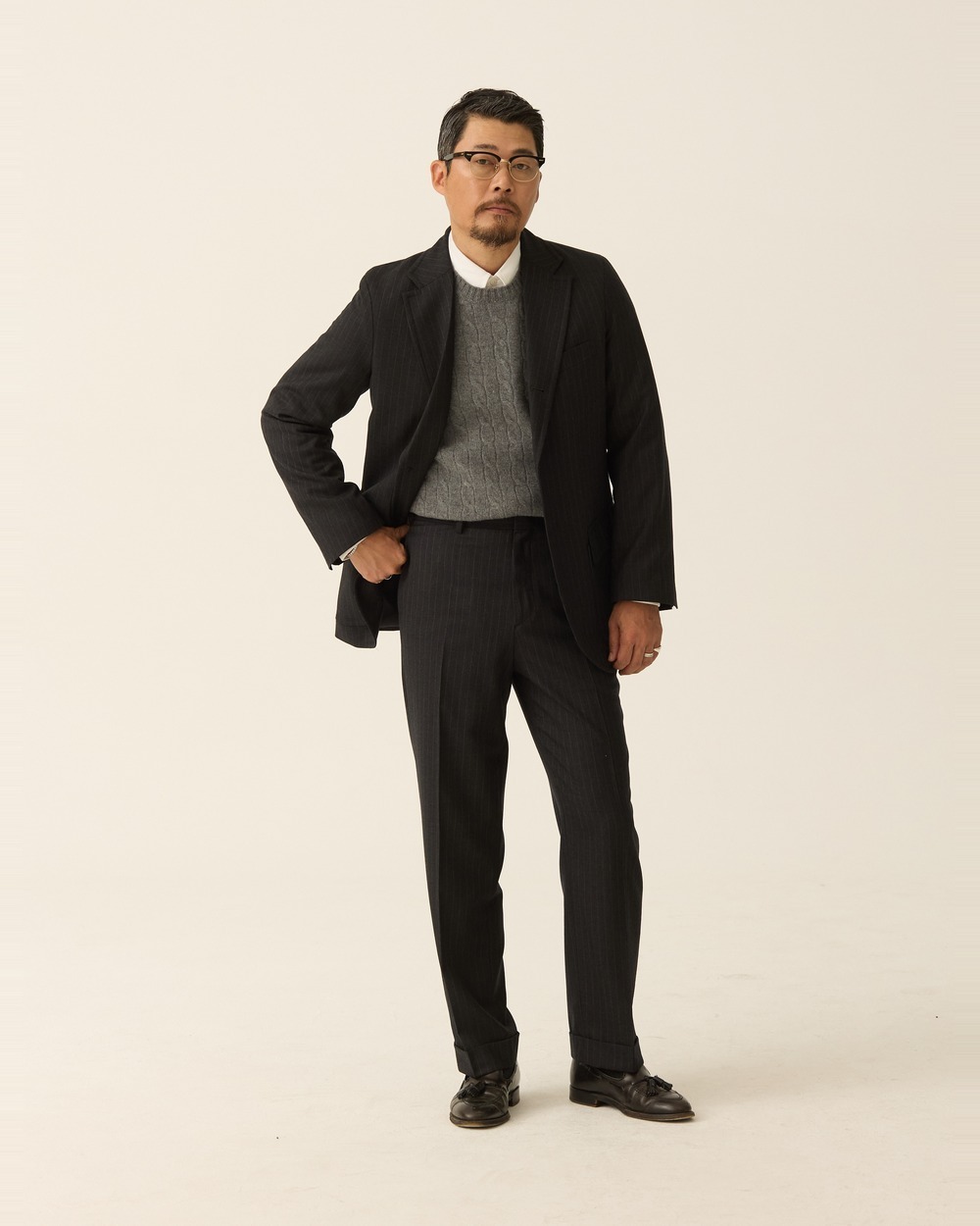 Full-length view of a man in a charcoal pinstripe sack suit worn open over a grey cable-knit crewneck sweater and a white shirt, no tie, one hand on his hip