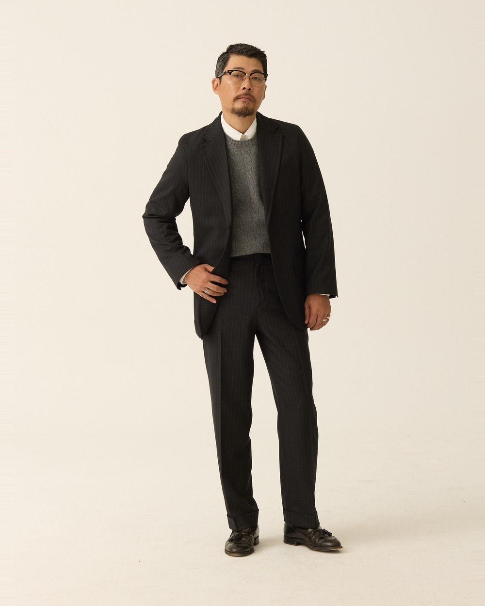 Full-length view of a charcoal pinstripe suit with a grey crewneck sweater and white shirt, jacket open and one hand on his hip, over flat-front pipe-stem trousers