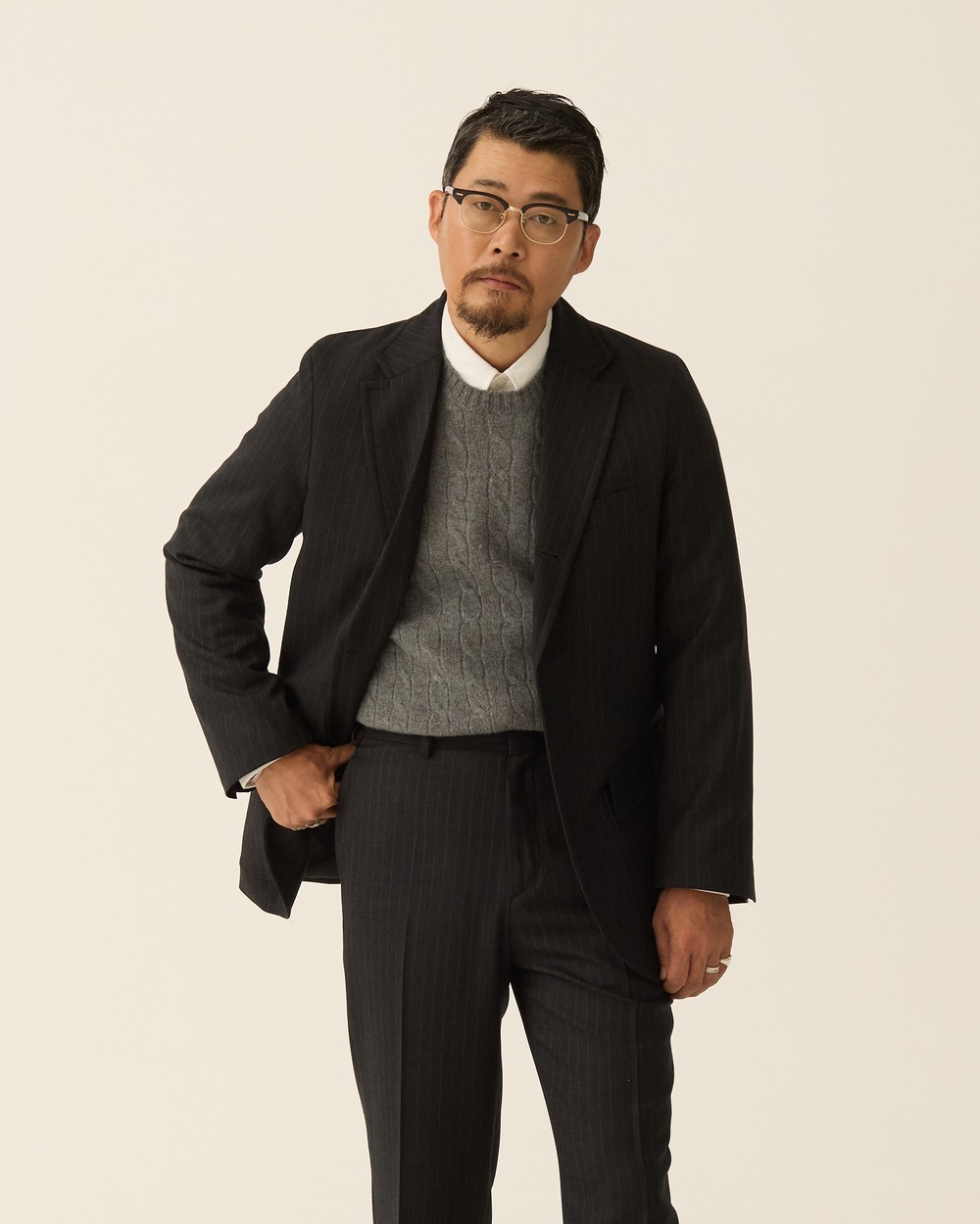 Man in a charcoal pinstripe sack jacket worn open over a grey cable-knit crewneck and a white shirt collar, one hand at his waist