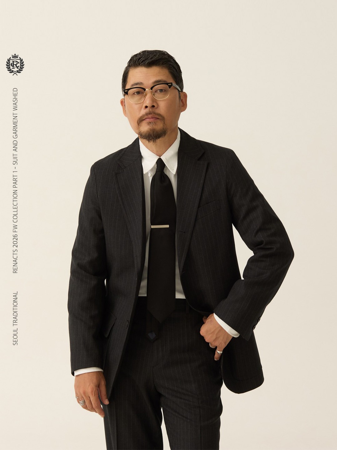 Man wearing a charcoal pinstripe sack suit with a white button-down shirt, collar points unbuttoned, a dark tie and a silver tie bar, one hand in his trouser pocket